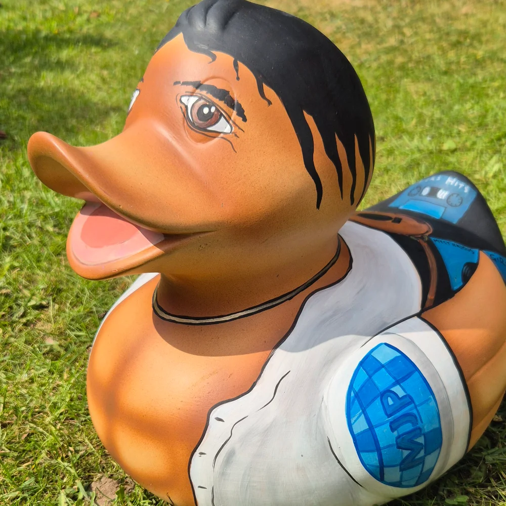 Close-up of a novelty rubber duck with stylised black hair, wearing a white vest and blue backpack, photographed outdoors on grass.