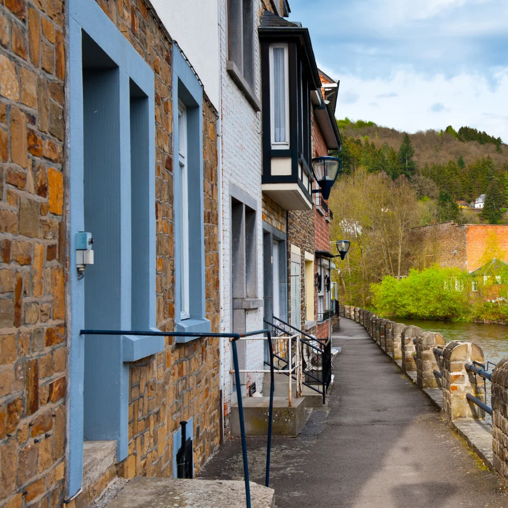 Stone houses beside a riverside embankment path with blue window surrounds, railings and wooded hillside views.